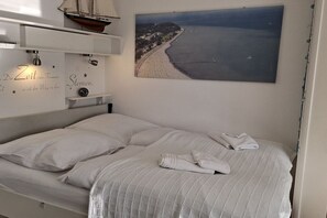Een netjes opgemaakt bed met een gewatteerde spreidek, een scheepsmodel op een plank erboven en een schilderij van een strand aan de muur.
