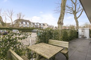 Ein Terrassenbereich mit einem Holztisch und einer Bank, umgeben von einem Metallzaun und Pflanzen.