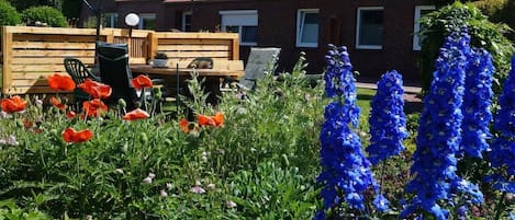 Un jardin avec des fleurs rouges et bleues, un banc en bois et un bâtiment en briques surmonté d’un toit en tuiles.
