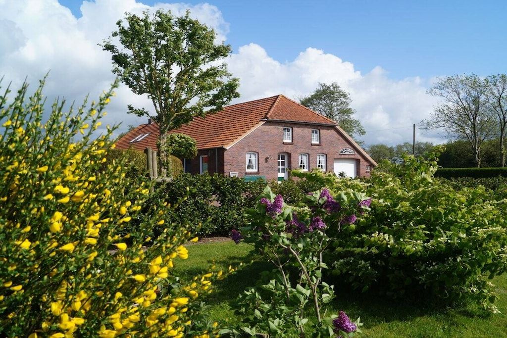 Una casa de dos pisos con techo de tejas rojas, rodeada de exuberante vegetación y plantas con flores.