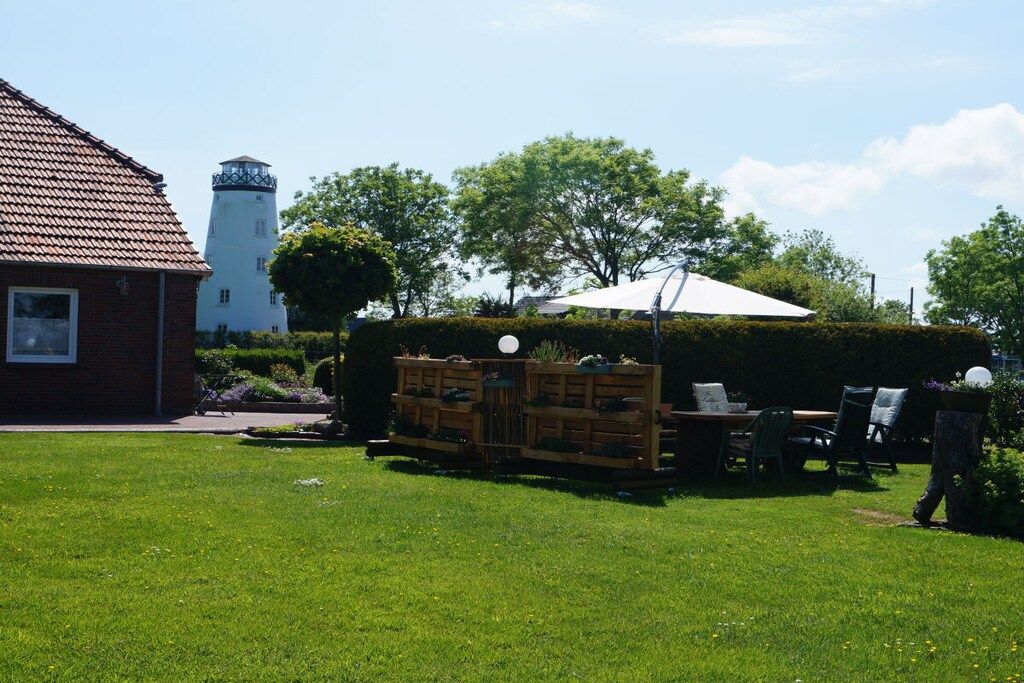 A garden with a wooden bench, a table, and chairs, a white umbrella, and a hedge.