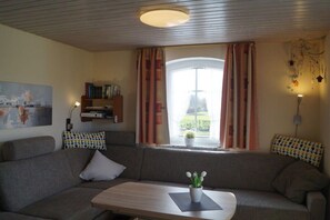 A living room with a sofa, a small table, a bookshelf, and a window with curtains. - Holiday apartment near the white tower (Ochtersum)