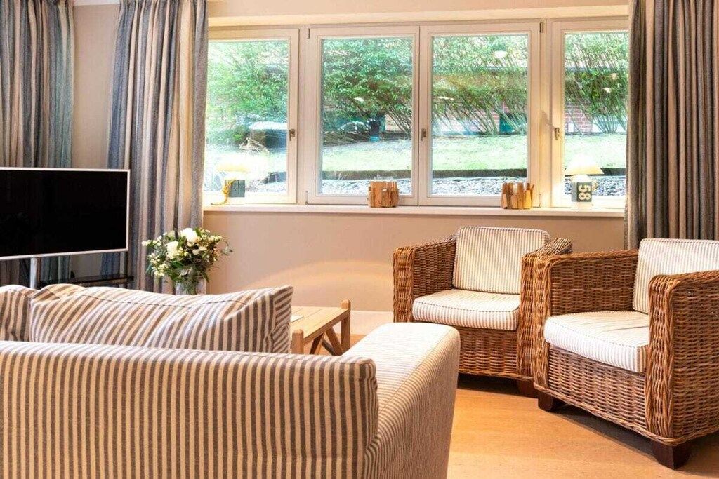 A room with a sofa, two wicker chairs, a TV, and a window with a view of greenery.