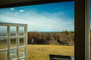 Blick durch ein Fenster auf einen wolkenlosen Himmel, eine Wiese und ein Gewässer in der Ferne.