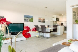 Ein modernes Wohnzimmer mit Flachbildfernseher, Essbereich mit Stühlen, einer Küche mit weißen Schränken und Kunstwerken an den Wänden.