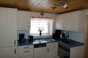 A kitchen with white cabinets, a blue countertop, a coffee maker, a sink, a stove, and a window with a curtain. - Mehnert's holiday apartment (Reinhardtsdorf)