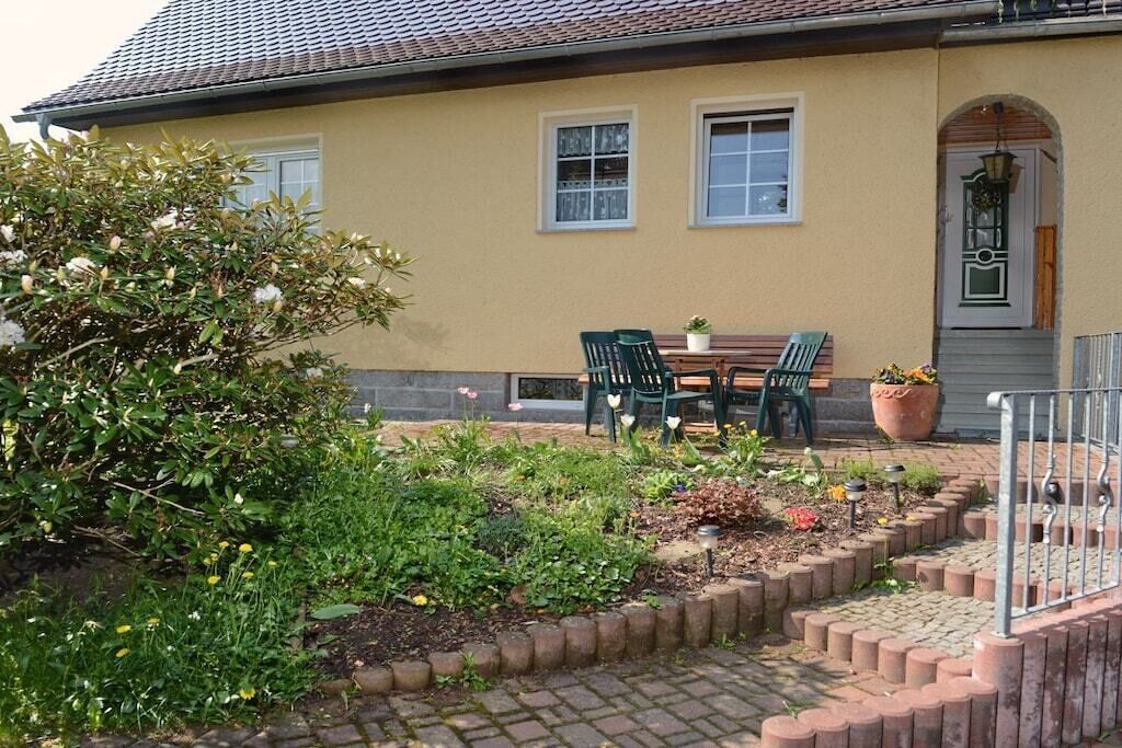 A yellow building with a tiled roof, a front door with a decorative arch, and a small garden area with a table and chairs.