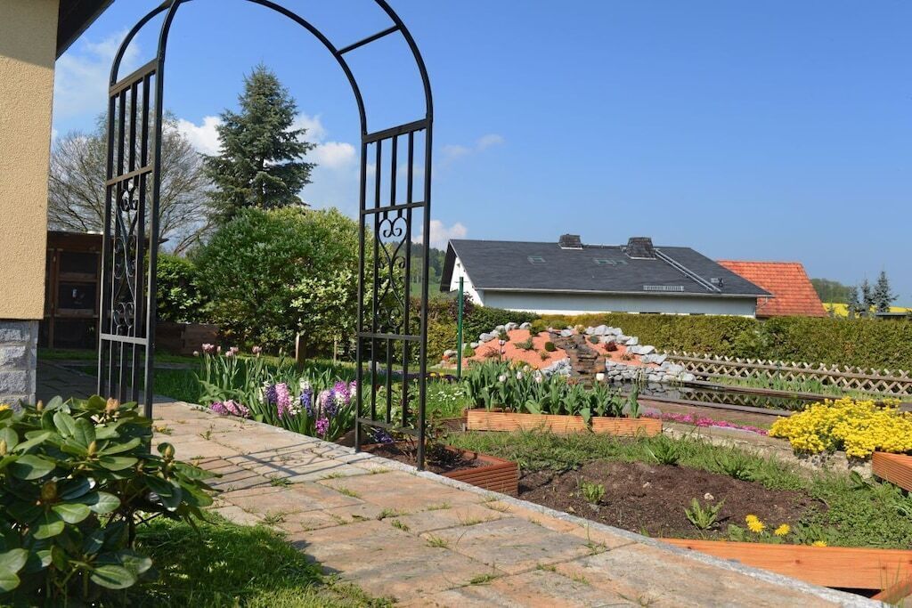 En have med et metalhegn, forskellige planter og et hus med rødt tag i baggrunden.
