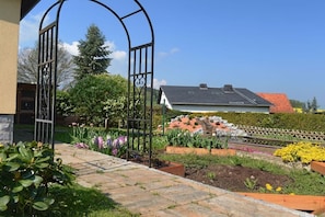 Ein Garten mit einem Metallzaun, verschiedenen Pflanzen und einem Haus mit rotem Dach im Hintergrund.