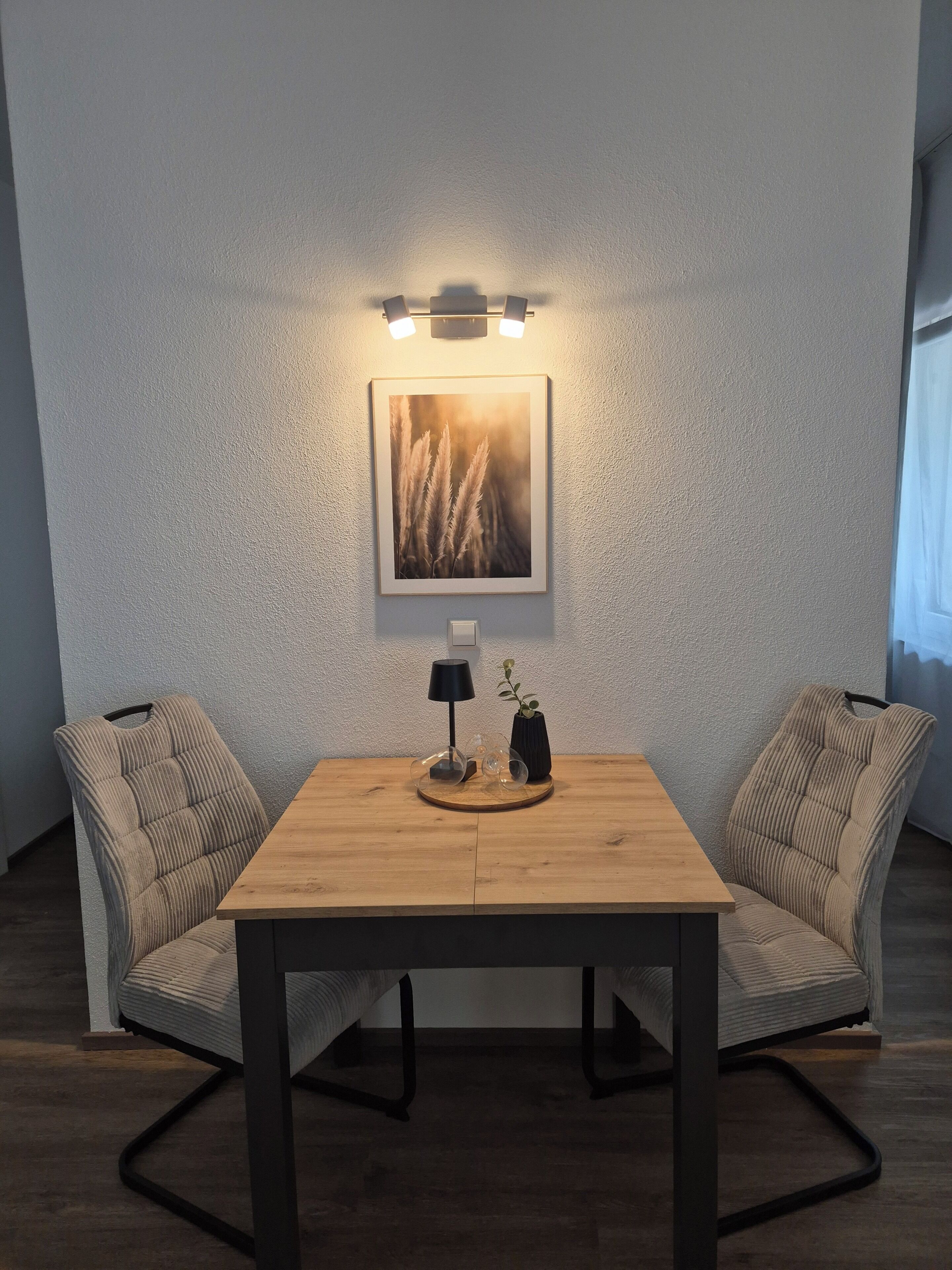 Un comedor con mesa de madera, dos sillas, una lámpara de pared y un cuadro enmarcado.