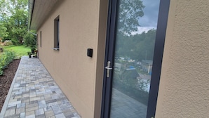 A building exterior with a paved walkway, a door with a glass panel, and a small garden area with plants.