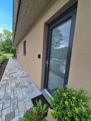 A building exterior with a paved walkway, a door with a glass panel, and a small garden area with plants.