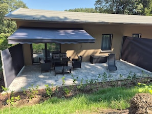 A covered patio with outdoor furniture, a window, and a view of the surrounding greenery.