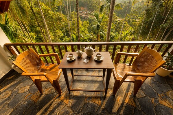 A balcony with two wooden chairs and a table, a potted plant, and a view of a lush green landscape.