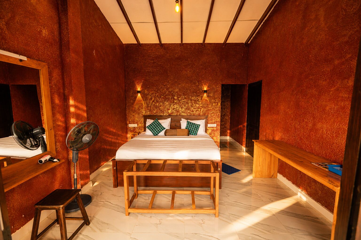 Un dormitorio con una cama de madera, un ventilador de pie, un banco y un taburete pequeño.