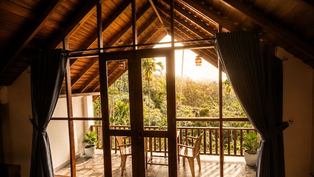 A room with a wooden ceiling, large windows with curtains, a view of a balcony with chairs and potted plants.