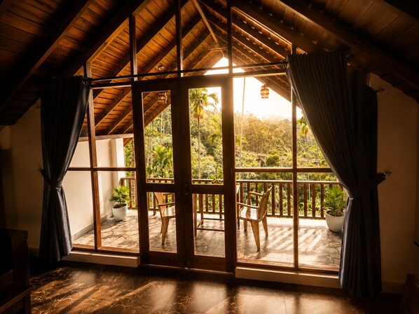 A room with a wooden ceiling, large windows with curtains, a view of a balcony with chairs and potted plants. - Nine Arch Jungle Inn (Ella)