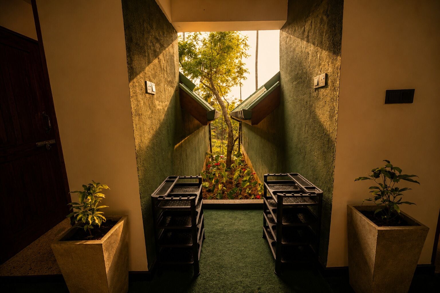 Un pasillo estrecho con vista a un jardín arbolado a través de una ventana, que conduce a una puerta. Hay dos macetas a cada lado del pasillo.
