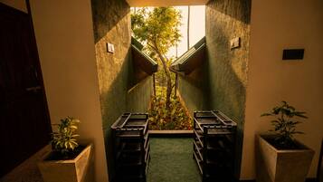 Un pasillo estrecho con vista a un jardín arbolado a través de una ventana, que conduce a una puerta. Hay dos macetas a cada lado del pasillo.