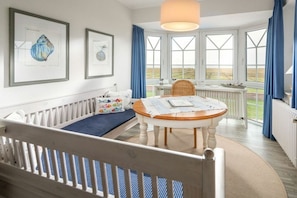 Ein Zimmer mit einem weißen Babybett, einem runden Holztisch und Blick durch große Fenster.