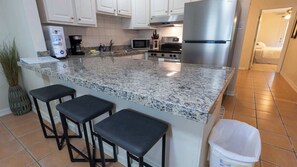 A kitchen with a granite countertop, stainless steel appliances, and three black bar stools. - 2BR Ground Floor, Sleeps 8, Pets ok - SPI (South Padre Island)