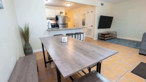 A dining area with a wooden table and benches, a kitchen in the background, and a television on the right. - 2BR Ground Floor, Sleeps 8, Pets ok - SPI (South Padre Island)