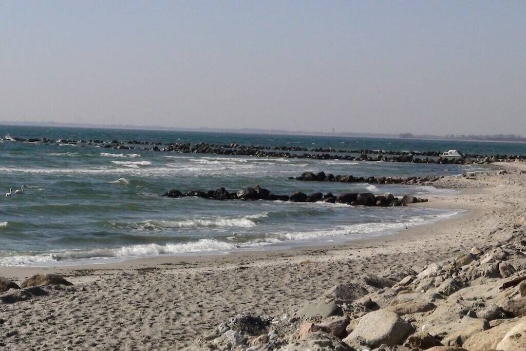 Ein felsiger Strand mit Wellen, blauem Himmel und einer fernen Stadtansicht.
