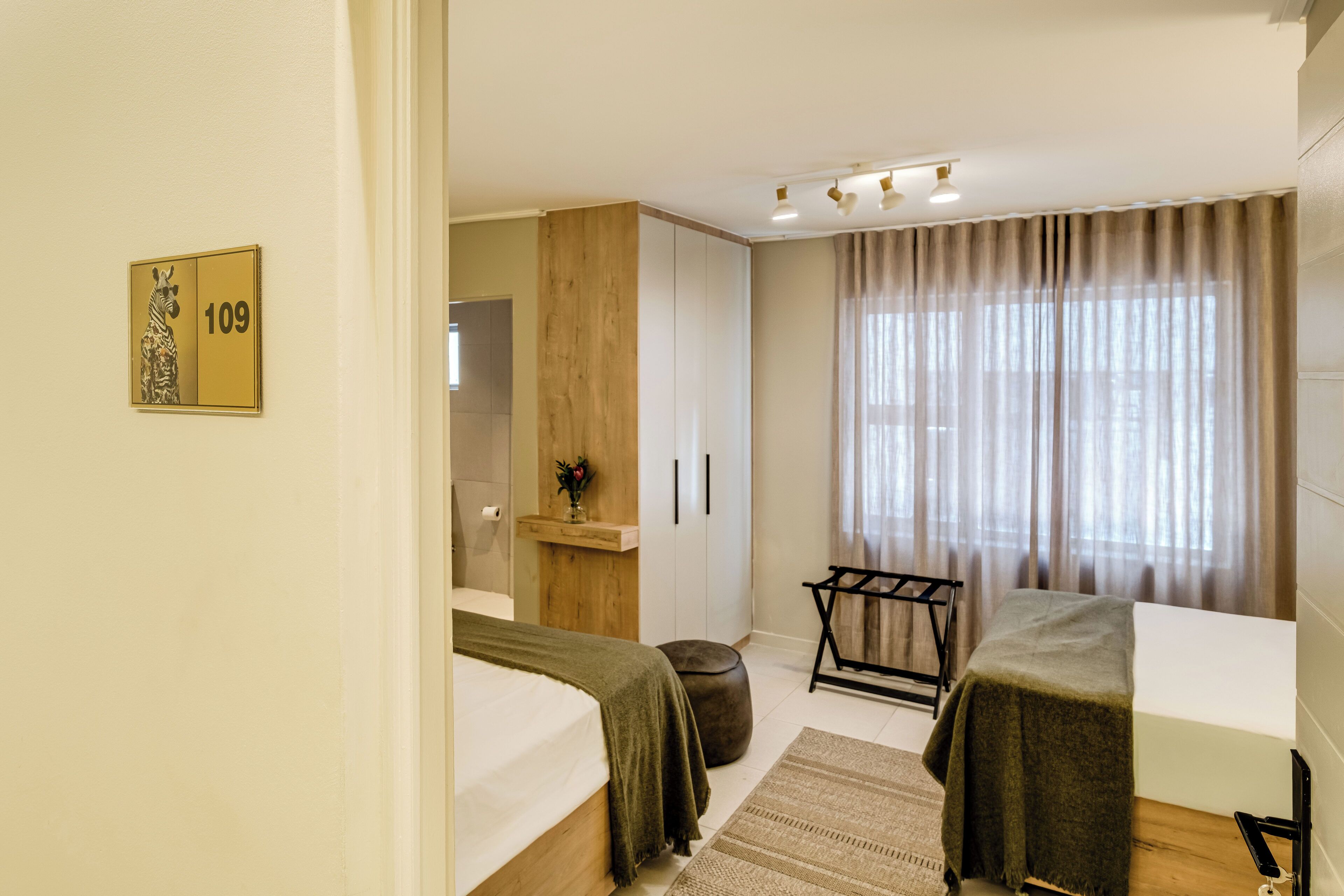 A hotel room with a bed, a desk, a chair, a window with curtains, and a wall-mounted number sign.