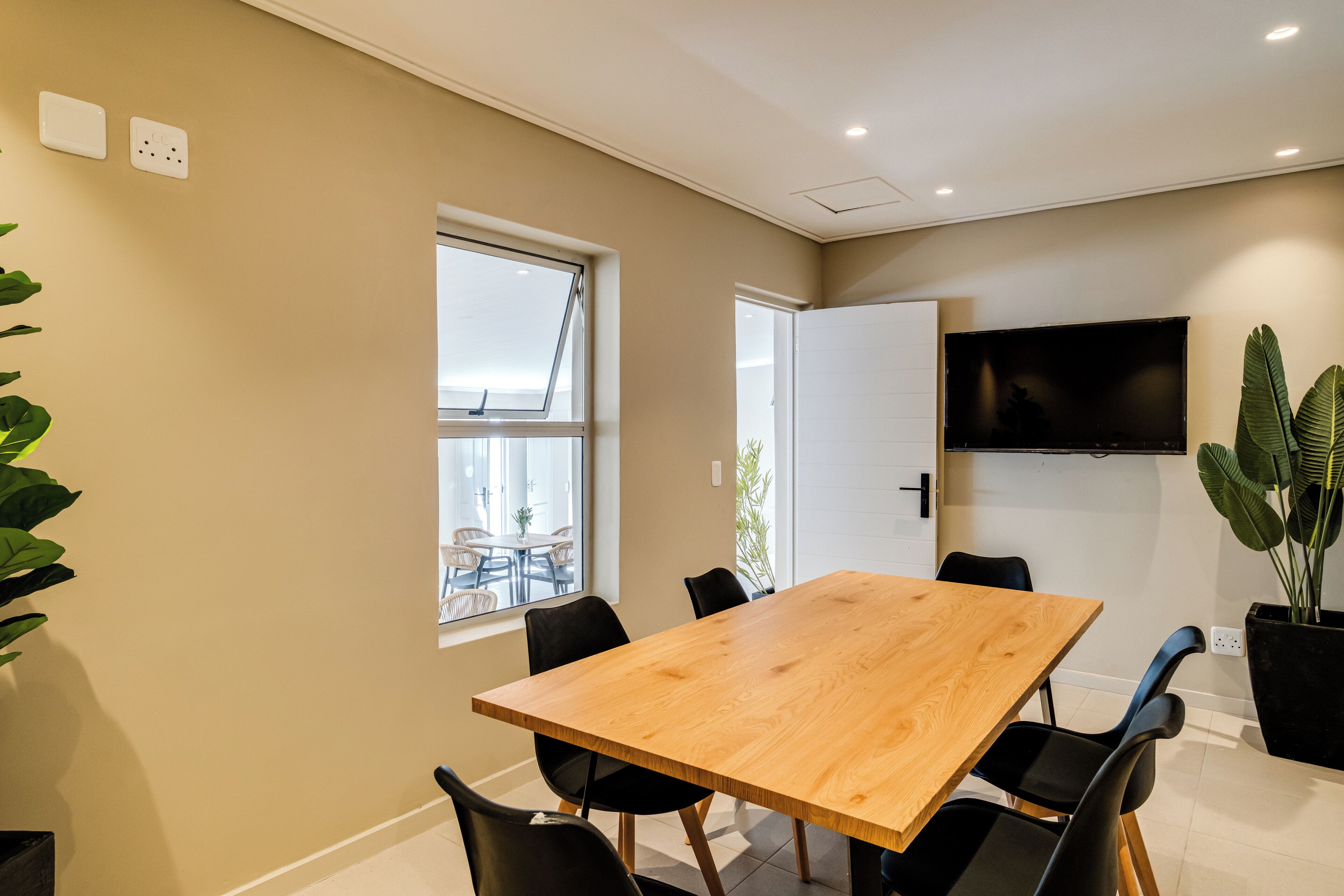 A modern meeting room with a wooden table, black chairs, a large window, and a flat-screen TV mounted on the wall.
