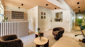 Un interior moderno con techo de listones de madera, una recepción, área de estar con sillas, una pequeña mesa redonda y una planta grande en maceta.