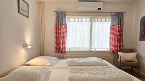 Une chambre à coucher avec un lit, deux oreillers, une chaise, une fenêtre avec des rideaux et un luminaire fixé au mur.