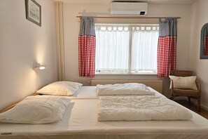 Un dormitorio con cama, dos almohadas, una silla, ventana con cortinas y una lámpara fijada a la pared.