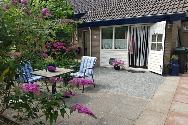 Un patio con mesa y sillas, una puerta con cortinas y un jardín lleno de flores rosas.