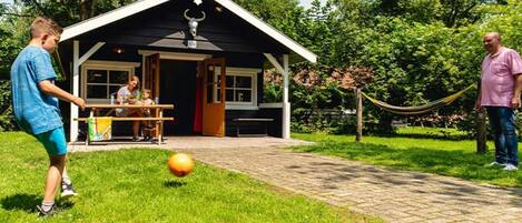A building with a covered patio area, a dining table with people seated, and a grassy area with a ball.