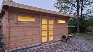 Eine Holzhütte mit einem kiengepflasterten Eingangsbereich, großen Fenstern und einem kleinen Garten mit Blumentöpfen.