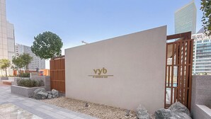 Um edifício moderno com fachada minimalista, um logotipo com as letras 'vyb' e o texto 'at business bay', e uma entrada gradeada com um caminho de cascalho e pedras.