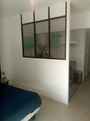 Quarto com cama, janela e uma kitchenette ao fundo.