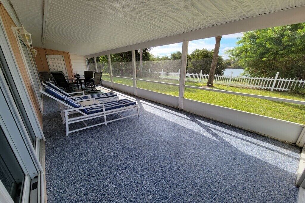 A covered outdoor area with lounge chairs and a table, large windows with a view of a grassy area and a fence.