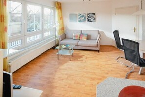 A living room with a sofa, a coffee table, and a chair. There are windows with curtains, a radiator, and artwork on the wall. - Cloverleaf in the Sanderling house (Juist)