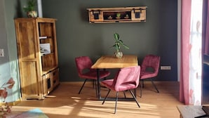 A room with a wooden dining table, two pink chairs, a wooden cabinet, and a shelf with plants. - Holiday apartment with 1 bedroom (Bayerisch Eisenstein)