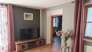 A living room with a television on a wooden stand, a small table with a vase of flowers, and a window with curtains. - Holiday apartment with 1 bedroom (Bayerisch Eisenstein)