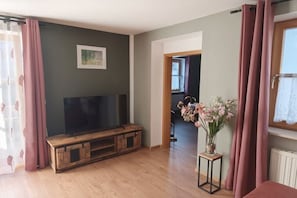 Ein Wohnzimmer mit Fernseher auf einem Holzständer, einem kleinen Tisch mit Blumenvase und einem Fenster mit Vorhängen.