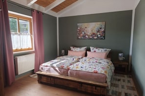 Ein Schlafzimmer mit hölzernem Bettgestell, floralem Bettbezug, einem Heizkörper, einem Fenster mit Vorhängen und einem Bild an der Wand.