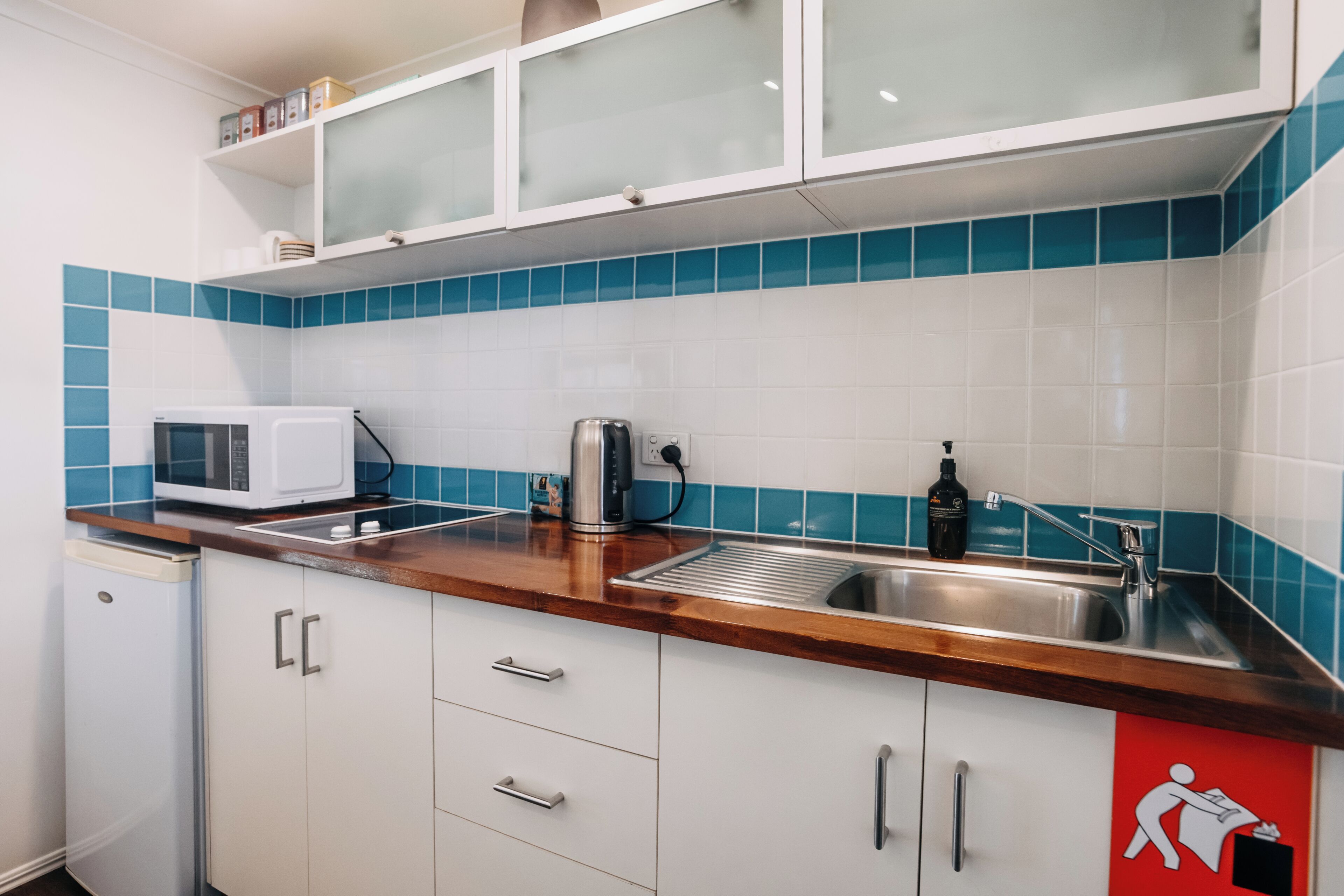 Una cocina moderna con armarios blancos, microondas, hervidor y pileta.