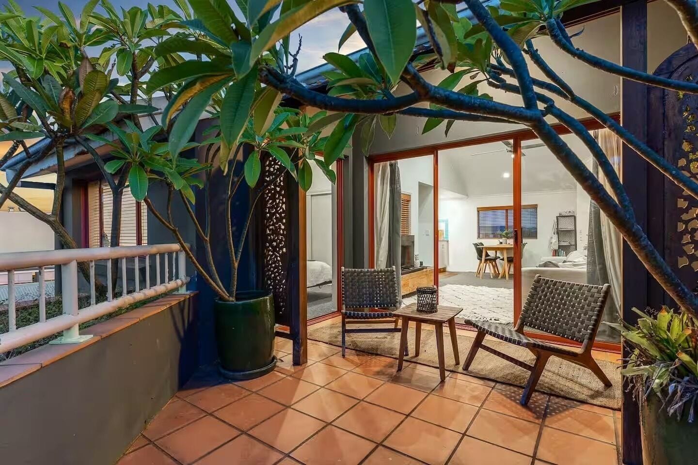 A balcony with a tiled floor, two wicker chairs, a potted plant, and a view into the interior of a house.