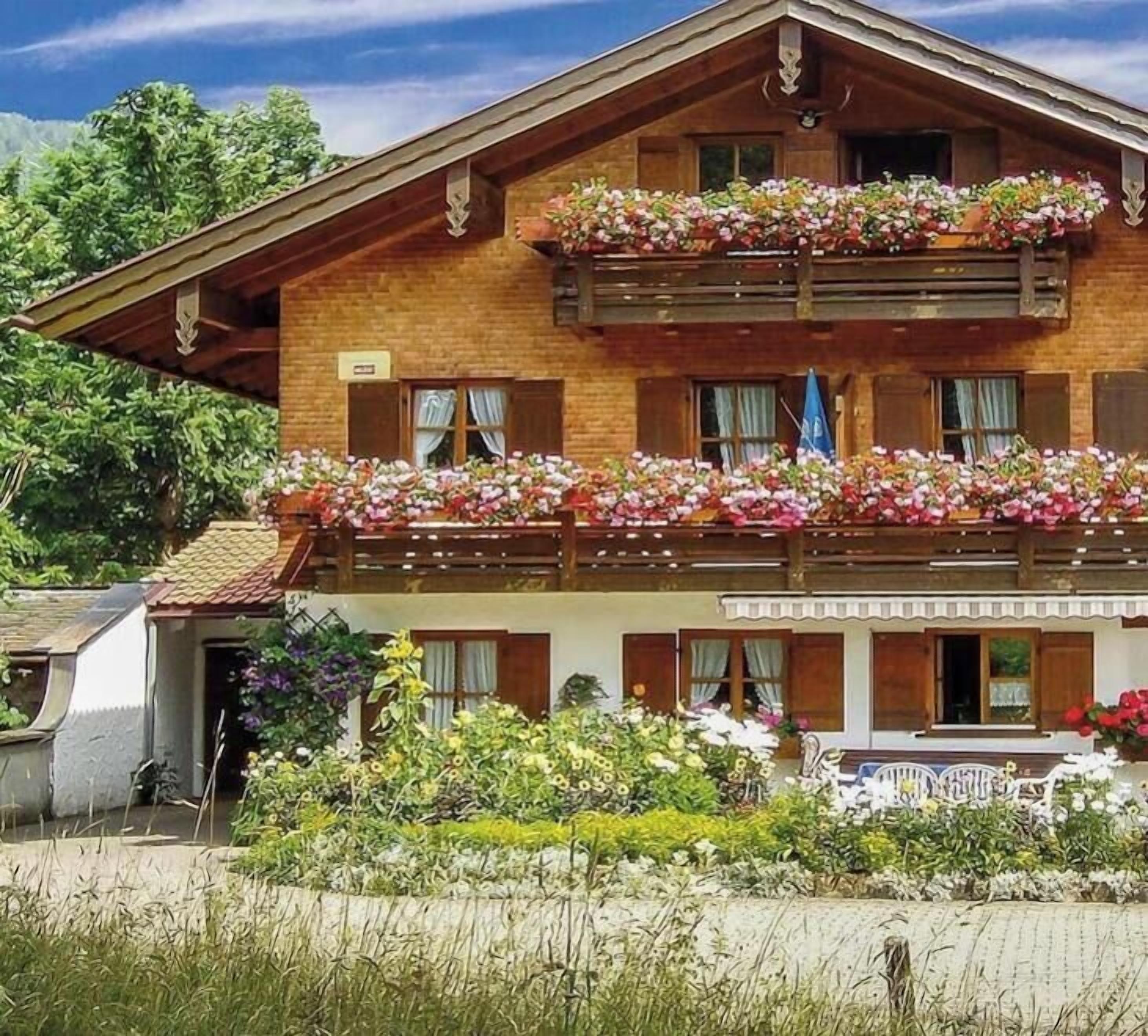 A two-story wooden building with balconies adorned with flowers, a covered entrance, and a garden area.