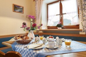 A breakfast setup with a checkered tablecloth, a vase of flowers, a basket of bread, and a window with curtains.