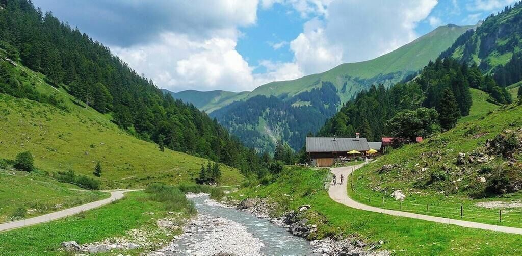 Un paysage de montagne avec une rivière, un sentier et un bâtiment doté d’un toit rouge.