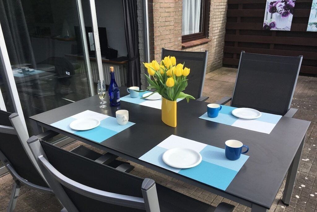 Un espace de restauration en plein air avec une table dressée pour deux personnes, agrémentée d’un vase de fleurs jaunes, d’une bouteille de vin et de sets de table bleus.