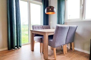 Un comedor con una mesa de madera y sillas grises tapizadas, una lámpara colgante y vistas a un paisaje verde a través de amplios ventanales.